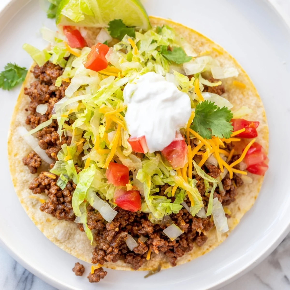 Ground beef simmered with cumin and chili powder sits inside soft flour tortillas, loaded with crisp lettuce, juicy tomatoes, shredded cheese, and a generous swirl of sour cream, garnished with cilantro and lime.