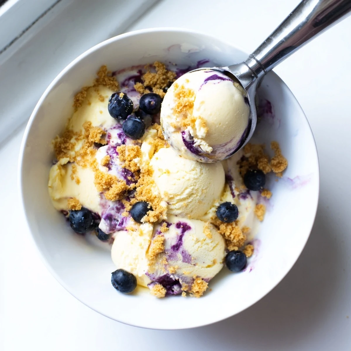 A scoop of Lemon Blueberry Cheesecake Ice Cream in a bowl with a fresh blueberry garnish and graham cracker crumbs.
