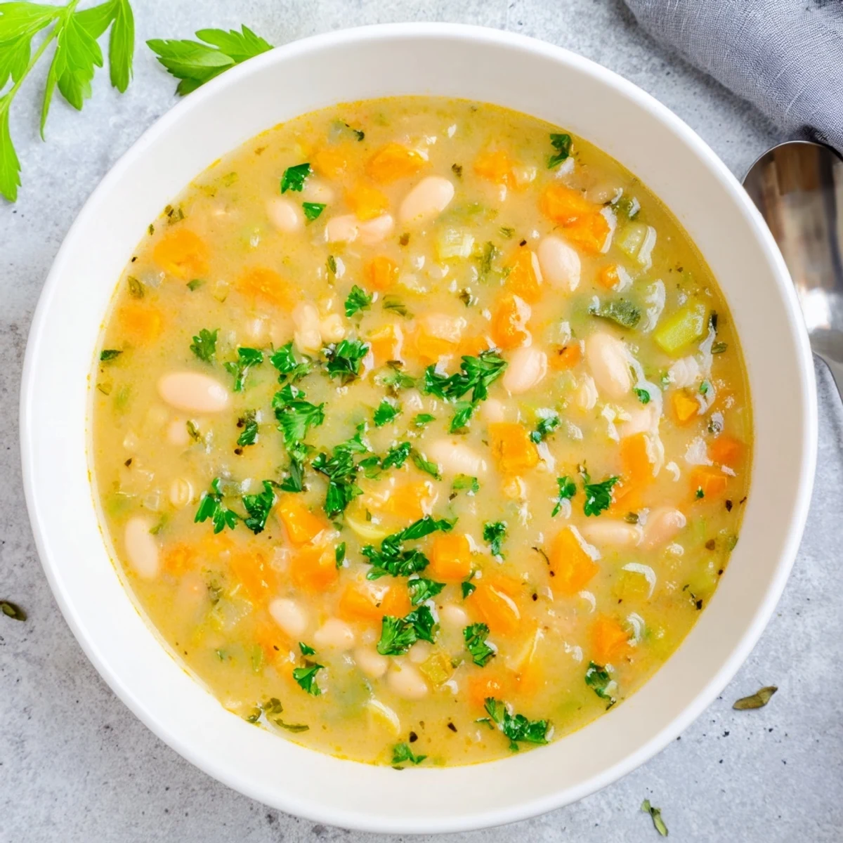 Rustic Mediterranean Cozy Rosemary Garlic White Bean Soup in a bowl beside olive oil, garlic, and fresh rosemary sprigs.