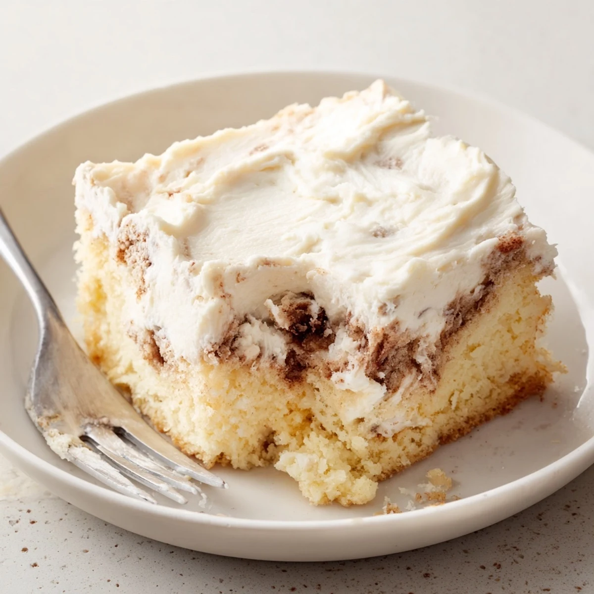 A fork beside Cinnamon Roll Poke Cake, cream cheese frosting glistening