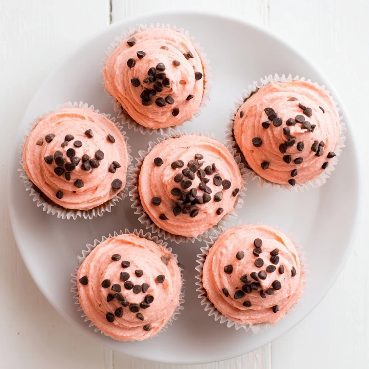 Fluffy pink-frosted watermelon cupcakes decorated with chocolate seeds on a white serving plate